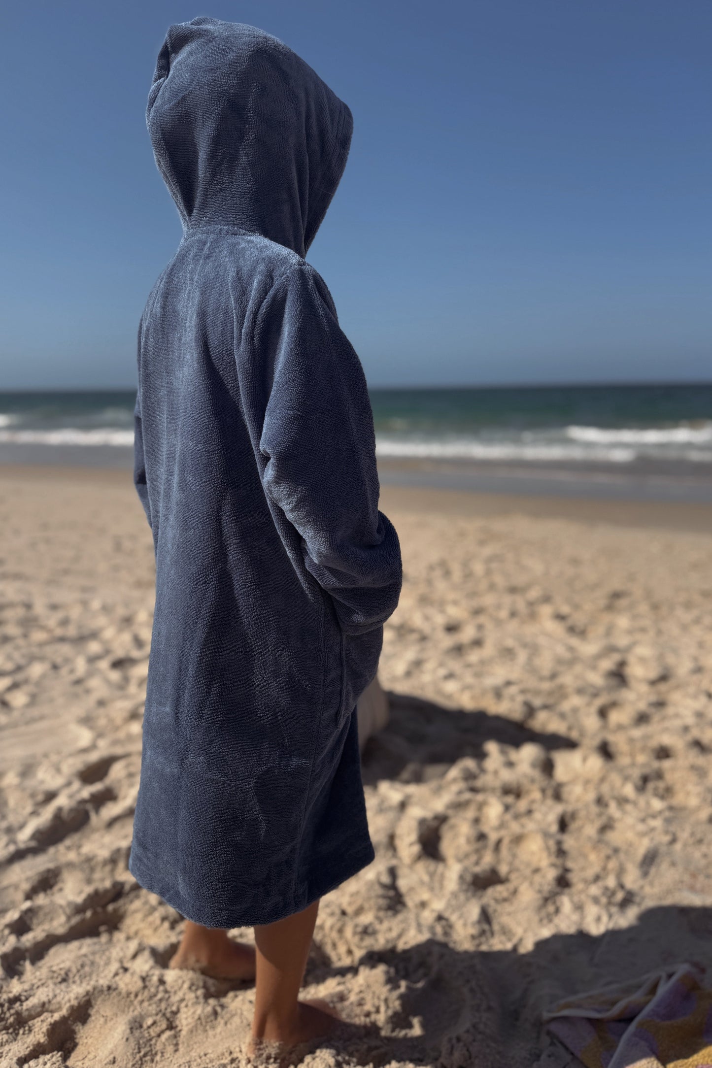 Kid wearing a blue hooded towel on a sandy beach with ocean waves in the background