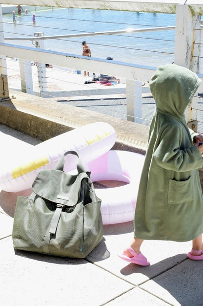 Person in a green robe standing next to a green bag on a tiled floor with a beach and pool area in the background.